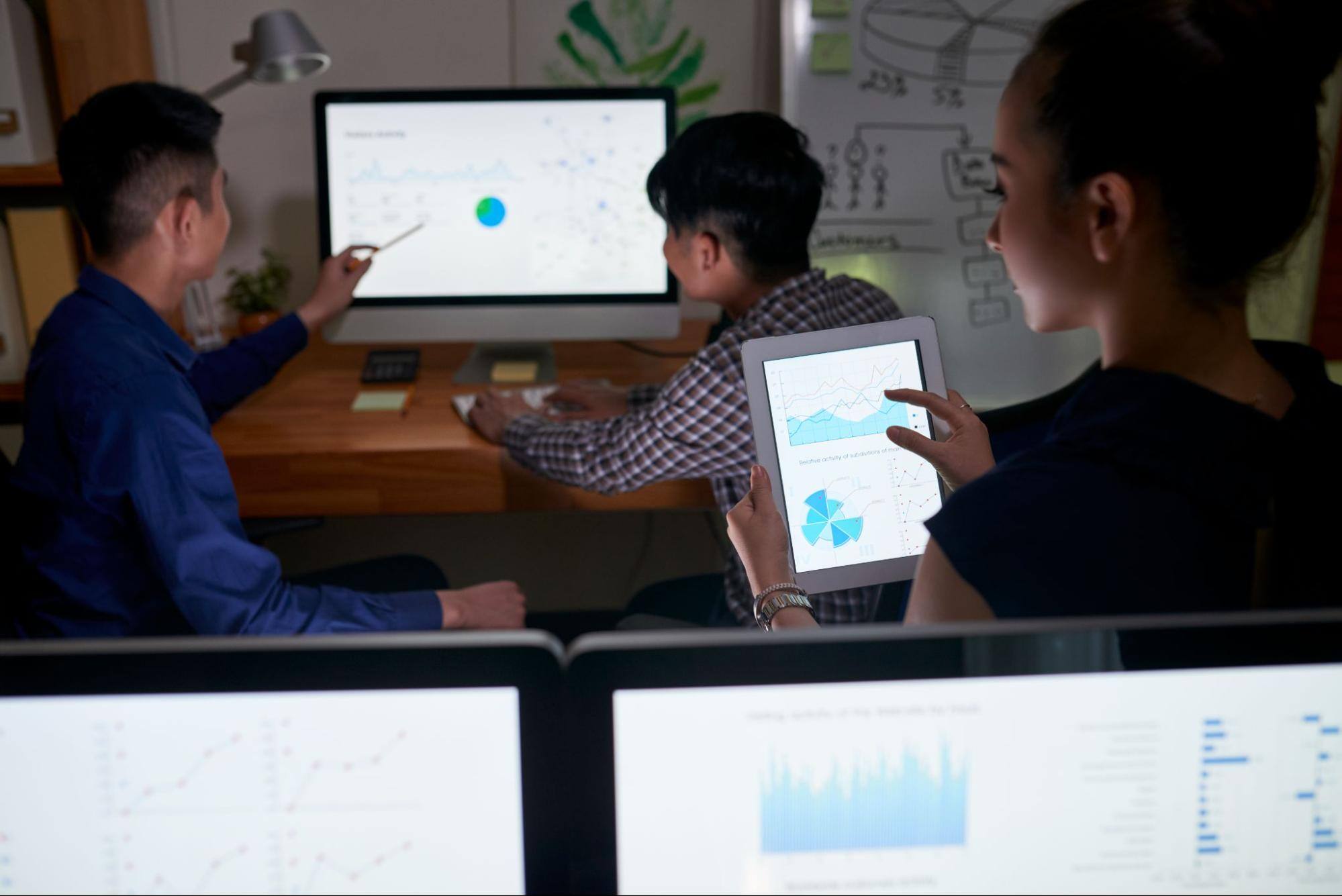 Two men are sitting down while looking at a computer screen, and a woman behind them is using a smart tablet.