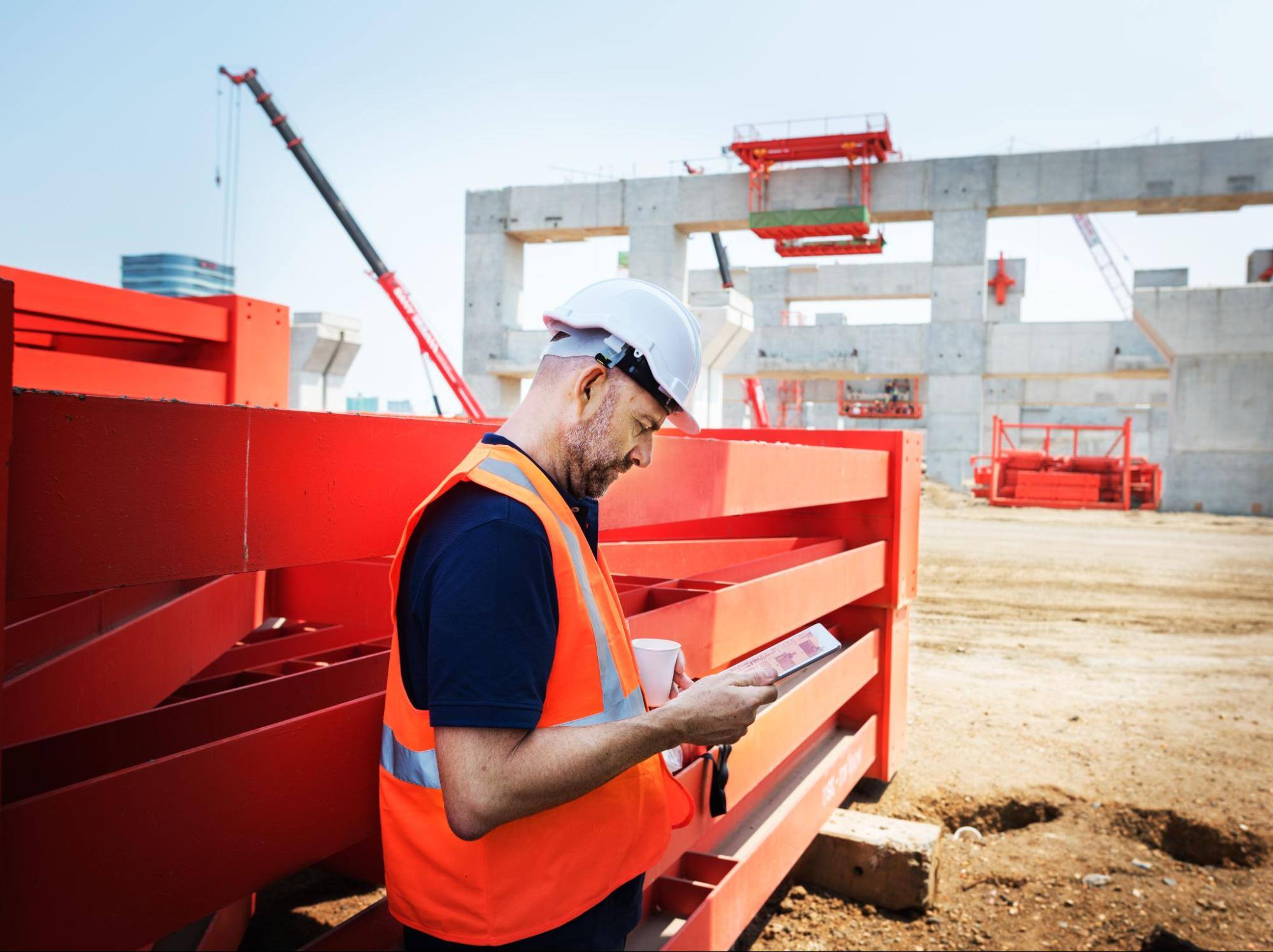 A worker wearing a caltrans orange vest and a hardhat glancing down at a smart tablet in the field at a construction site.