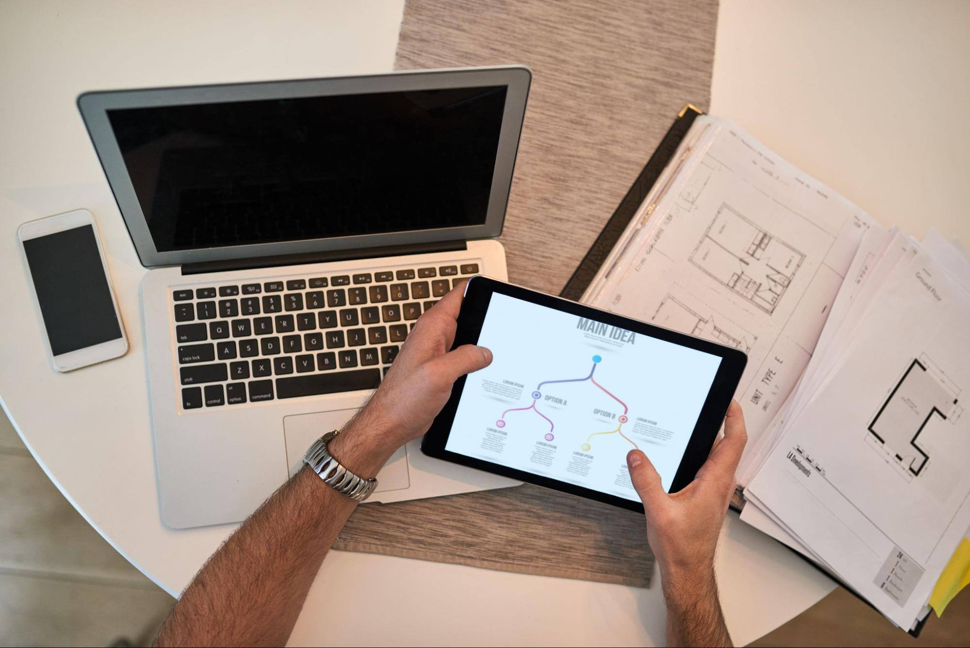 A view from above of a man’s hands holding a smart tablet with a laptop, a phone, and other documents surrounding him.