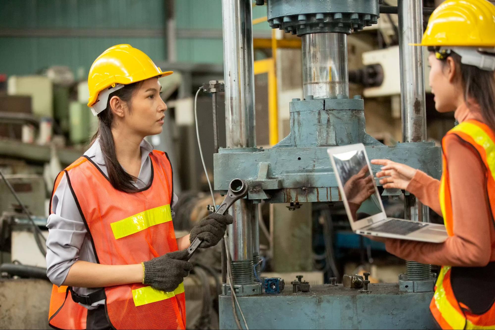 Two female construction workers with one holding a tool and the other holding a laptop. 