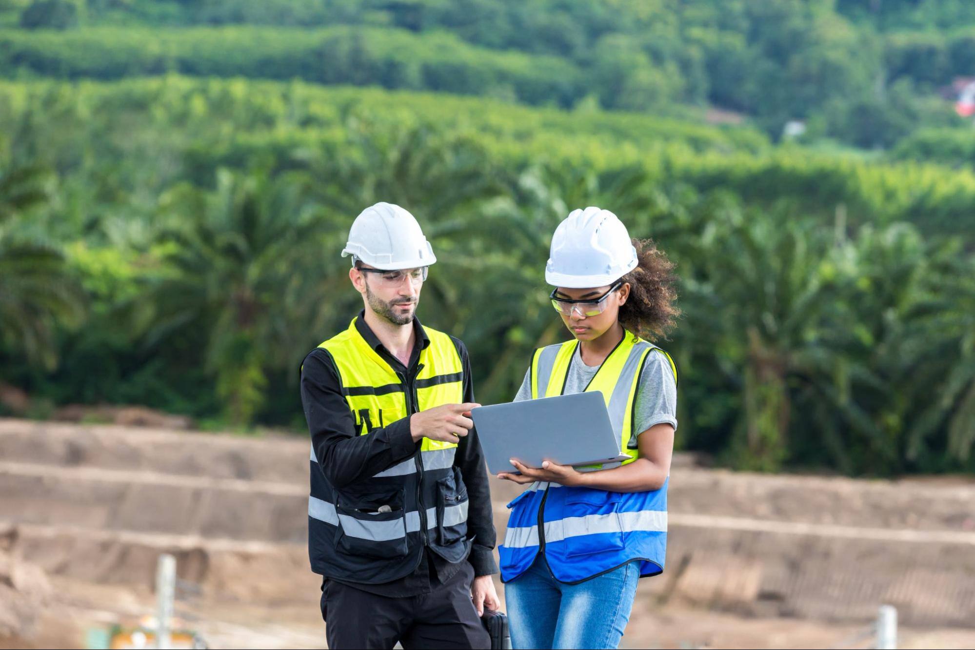 Two workers in hard hats looking at a computer outside. 