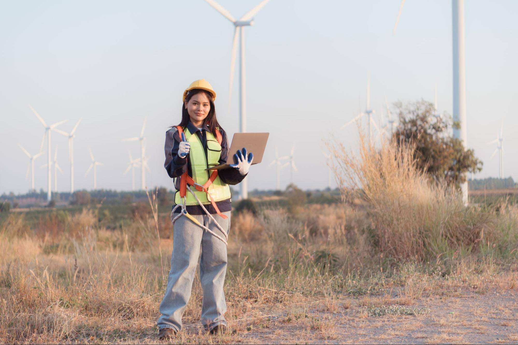 In a field, a woman with brown hair, wearing a uniform and a yellow hard hat, is holding a laptop and looking towards the camera.