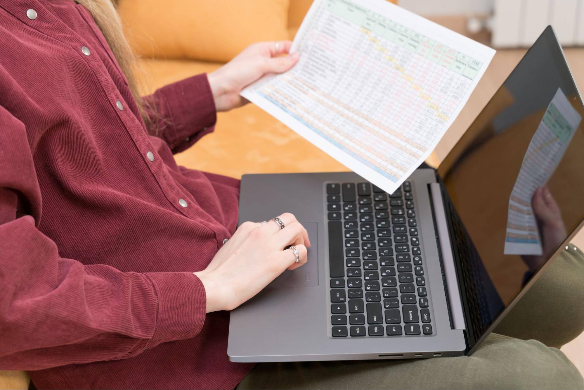 Cropped view of a woman holding a form in one hand and typing something into a laptop with the other hand.