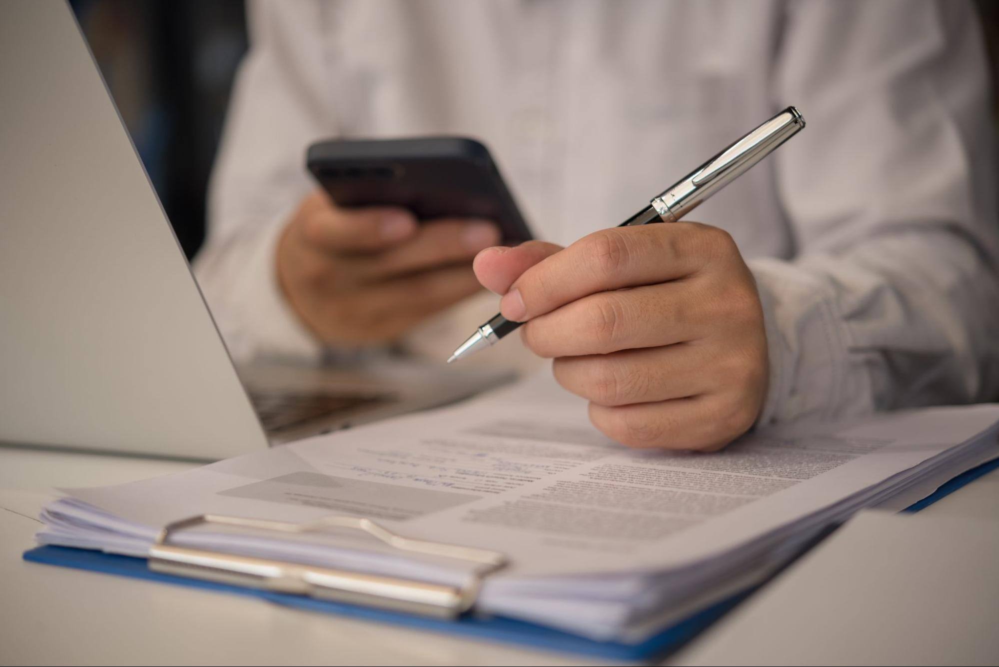 Cropped view of a man sitting behind a desk with a laptop in front of him, holding a pen over a paper form and his phone in the other hand.