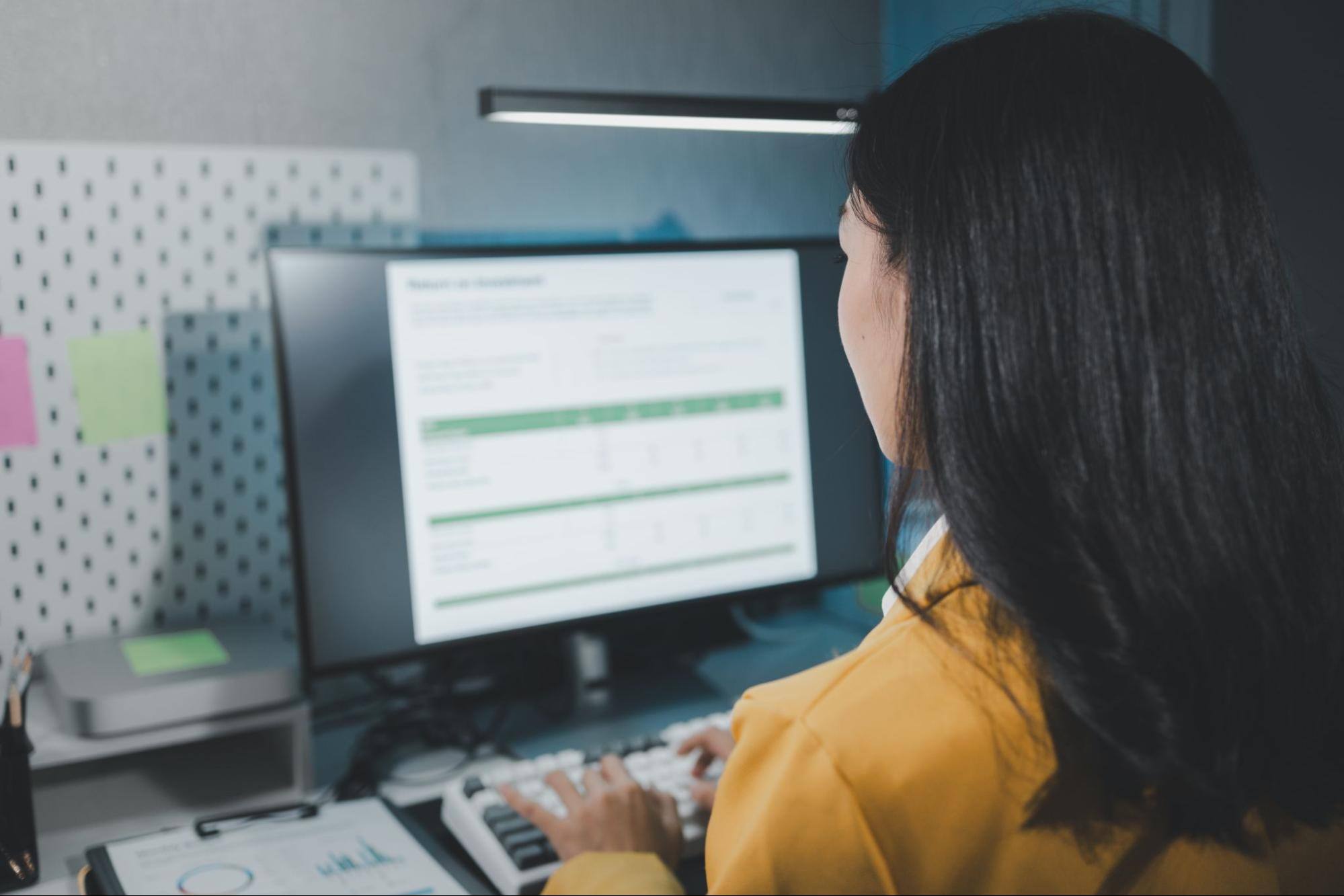 View of a woman with long brown hair wearing a yellow jacket typing information into a digital form on her computer.