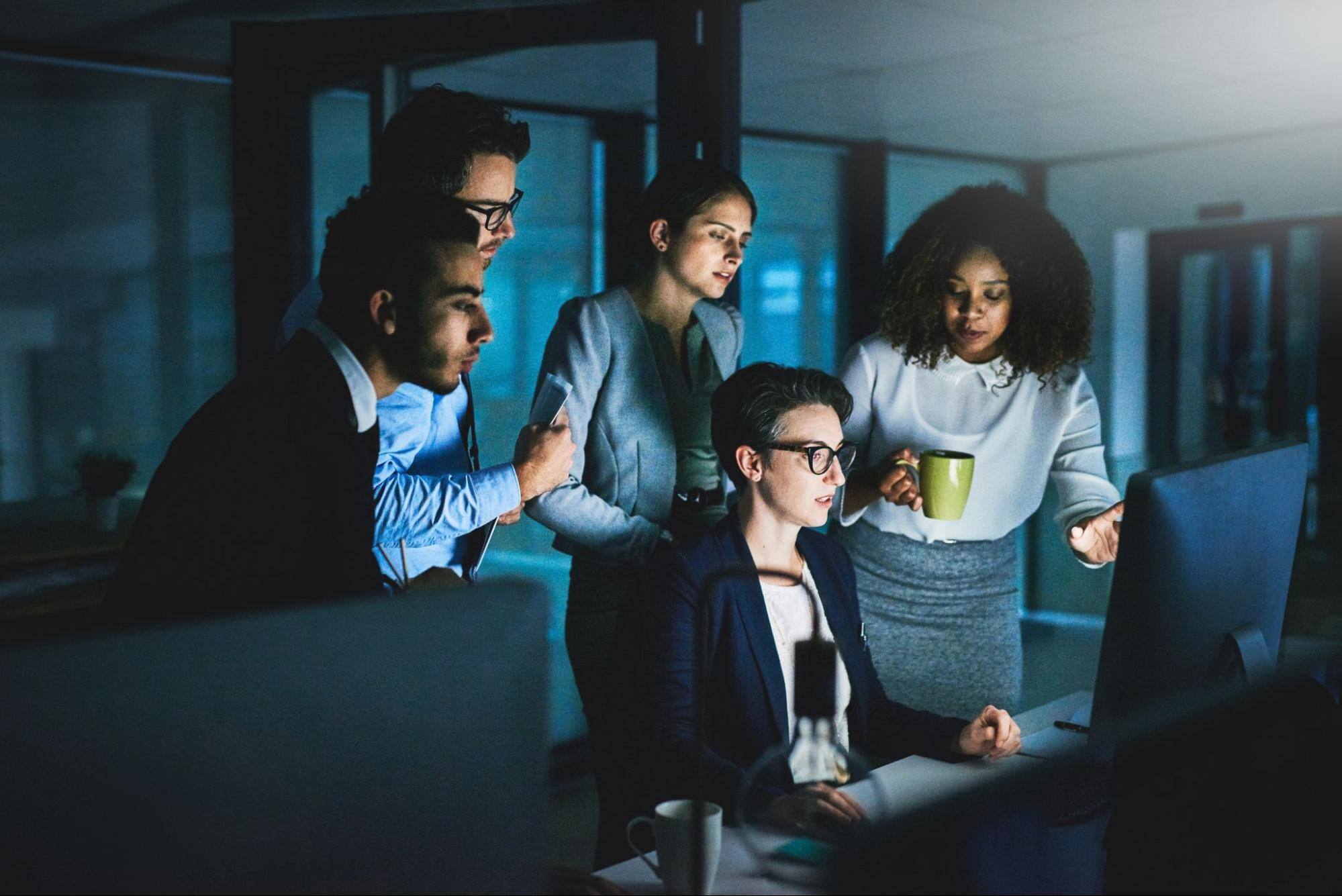 Five business people in an office, all looking at the same computer.