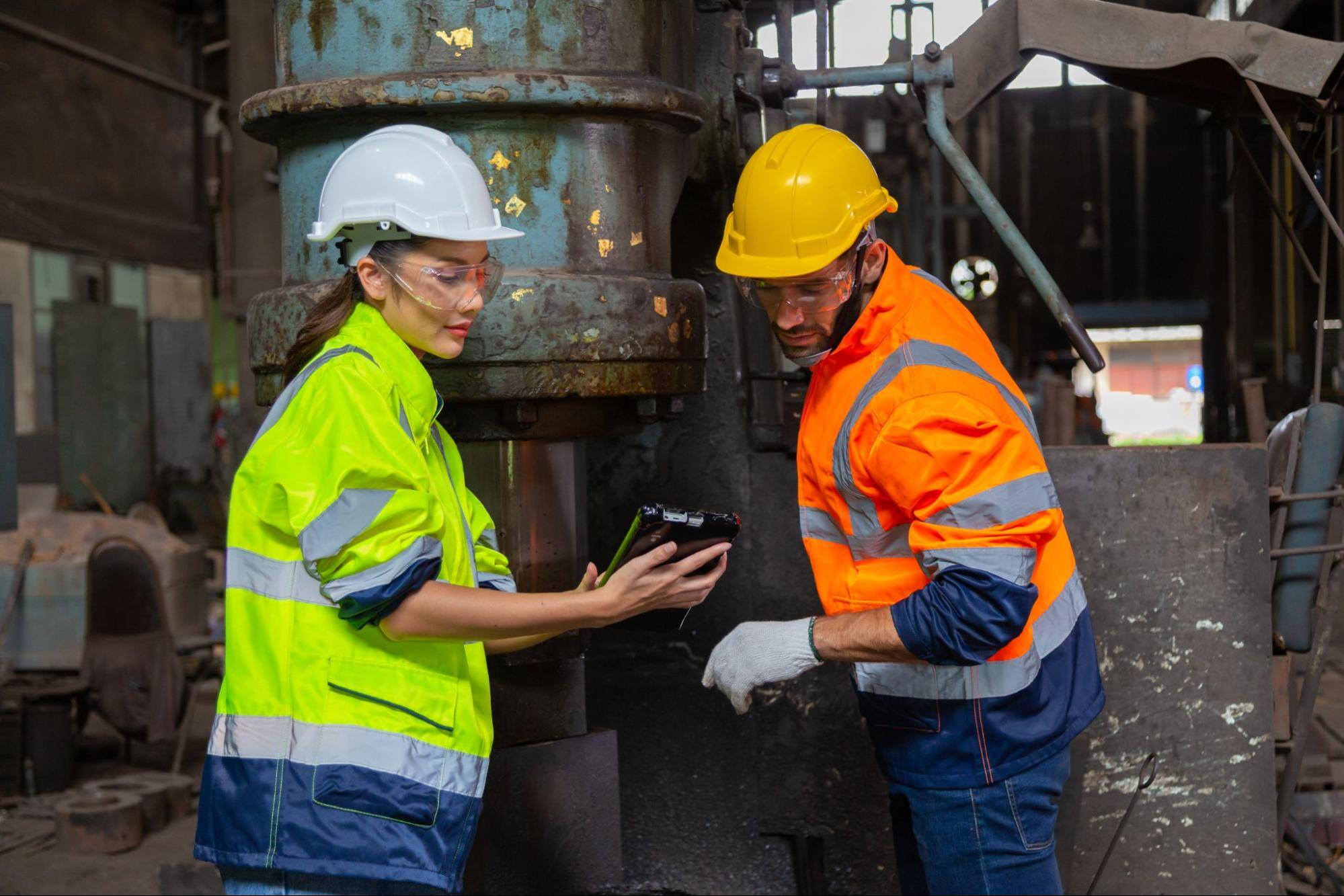 Two construction workers wearing hard hats and goggles looking at a tablet. 