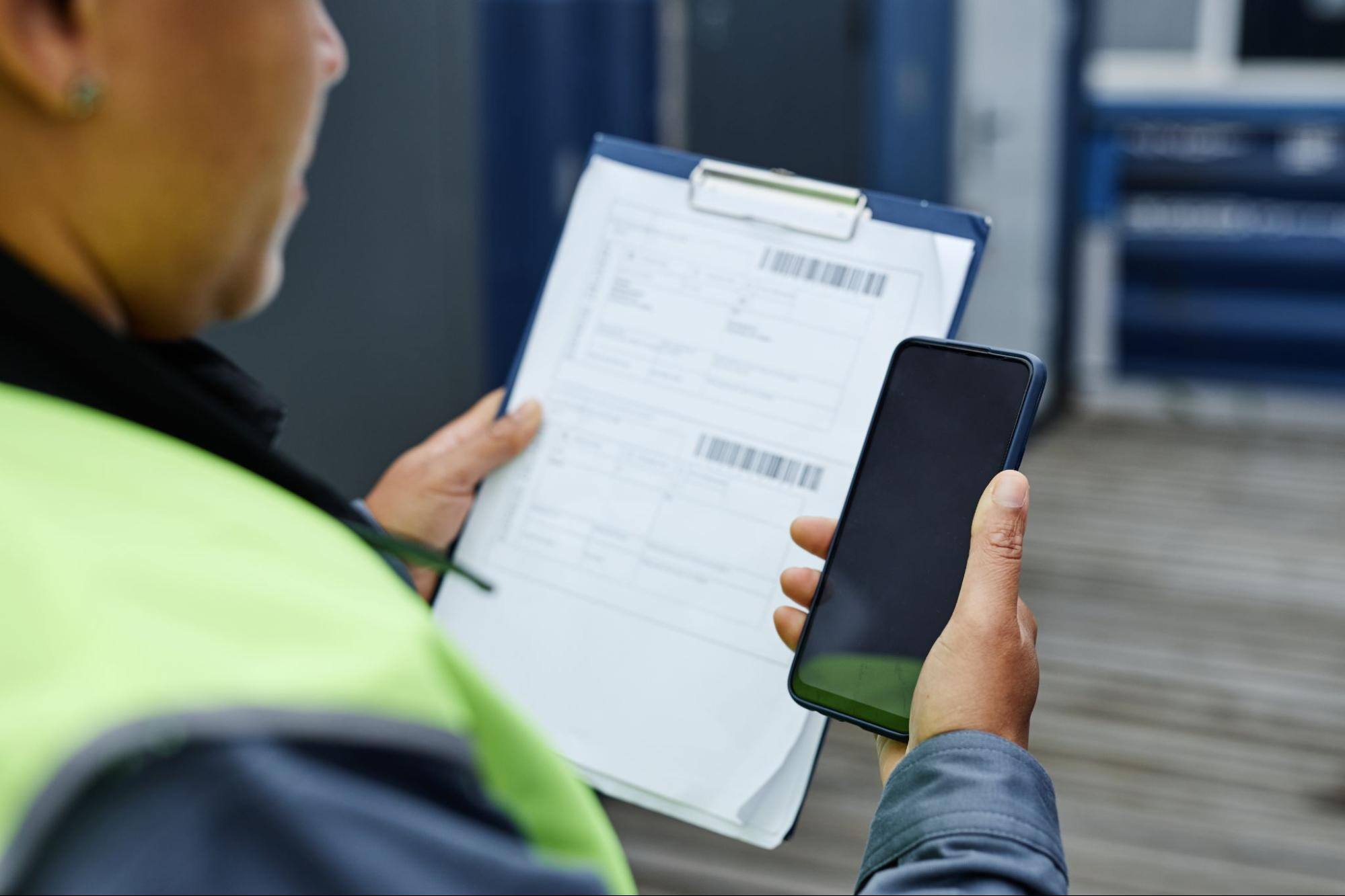 A cropped view of a worker glancing down while holding a paper form and a clipboard in one hand and a smartphone in the other.