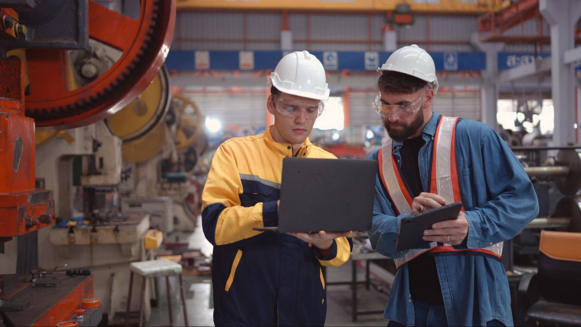 Two men wearing hard hats are in an industrial factory. One of the men is using a laptop, while the other is using a smart tablet.