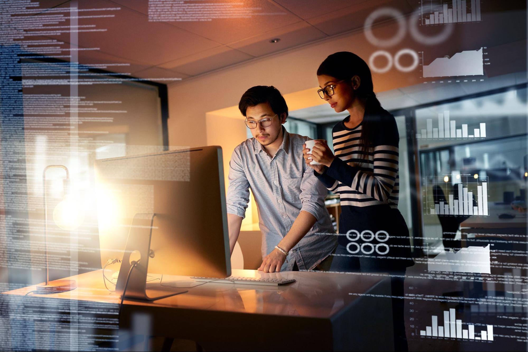 A man and a woman wearing glasses are standing while looking at a large computer screen with data imagery photoshopped on the photo.