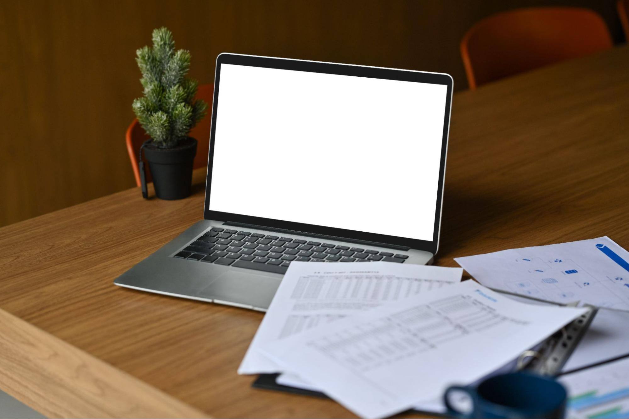 An open laptop and paper form next to each other on a wooden desk with a small potted plant in the background.