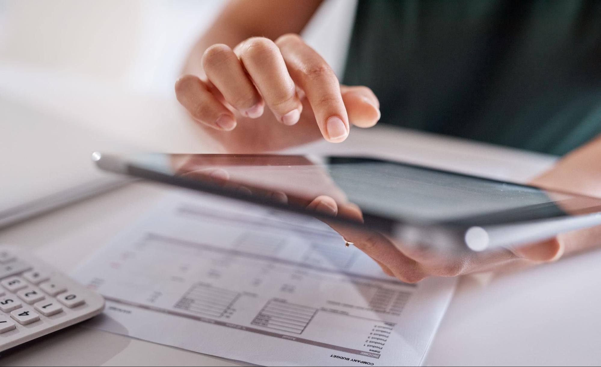 A cropped close-up view of a woman holding a smart tablet in one hand and using the other to input information with a paper form on the table in front of her.