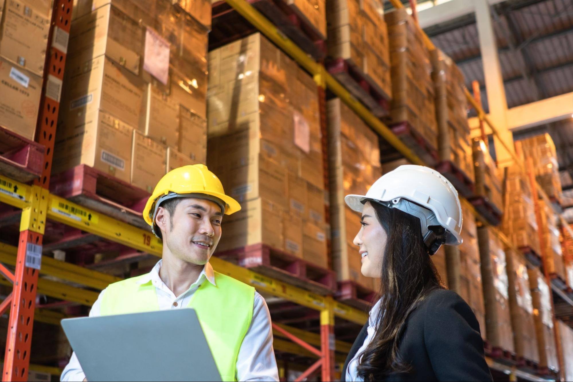 A man holding a laptop and a woman standing beside each other, smiling at each other in a warehouse with boxes surrounding them.