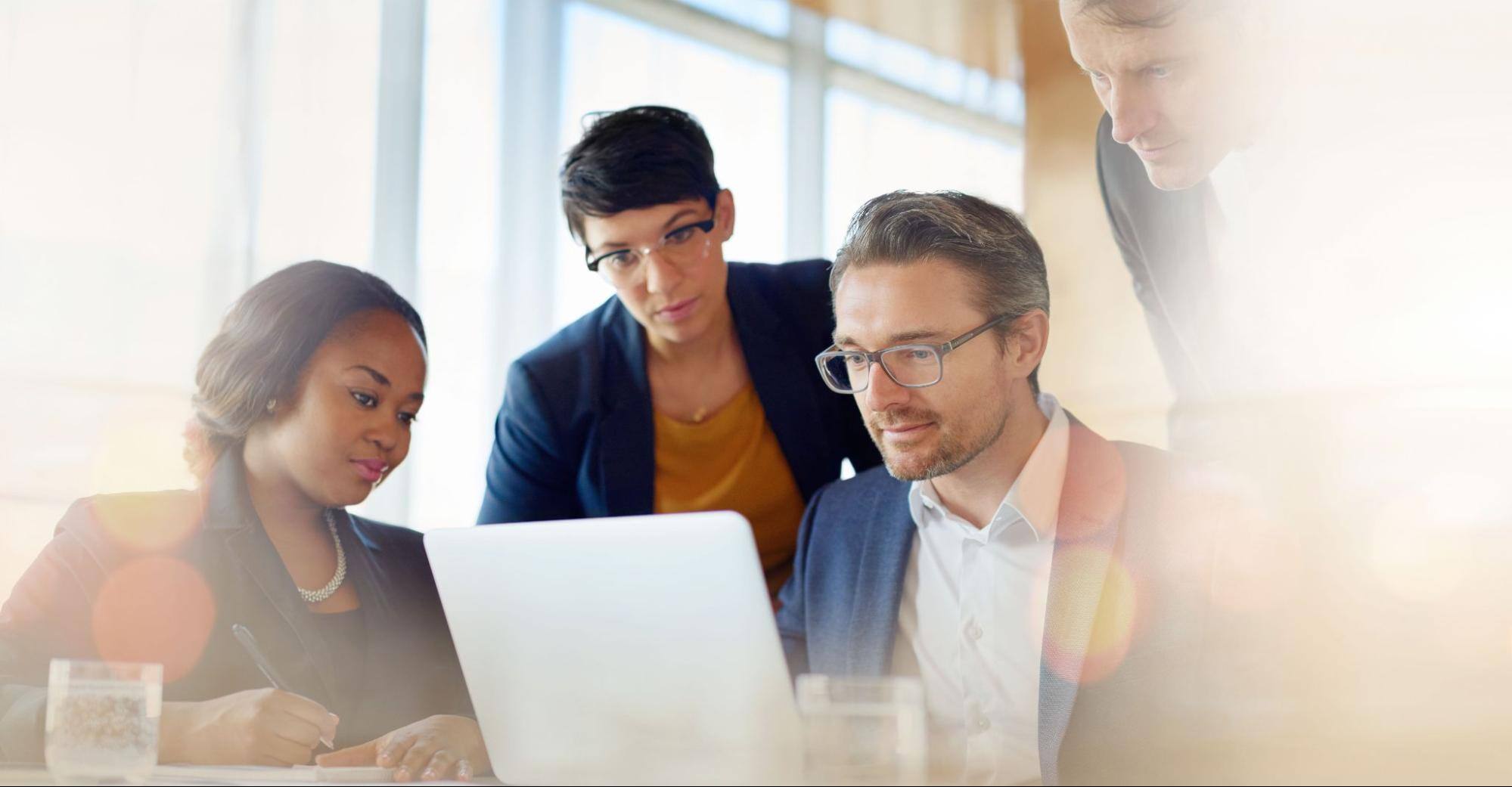 Four business people in an office, all looking at the same laptop.