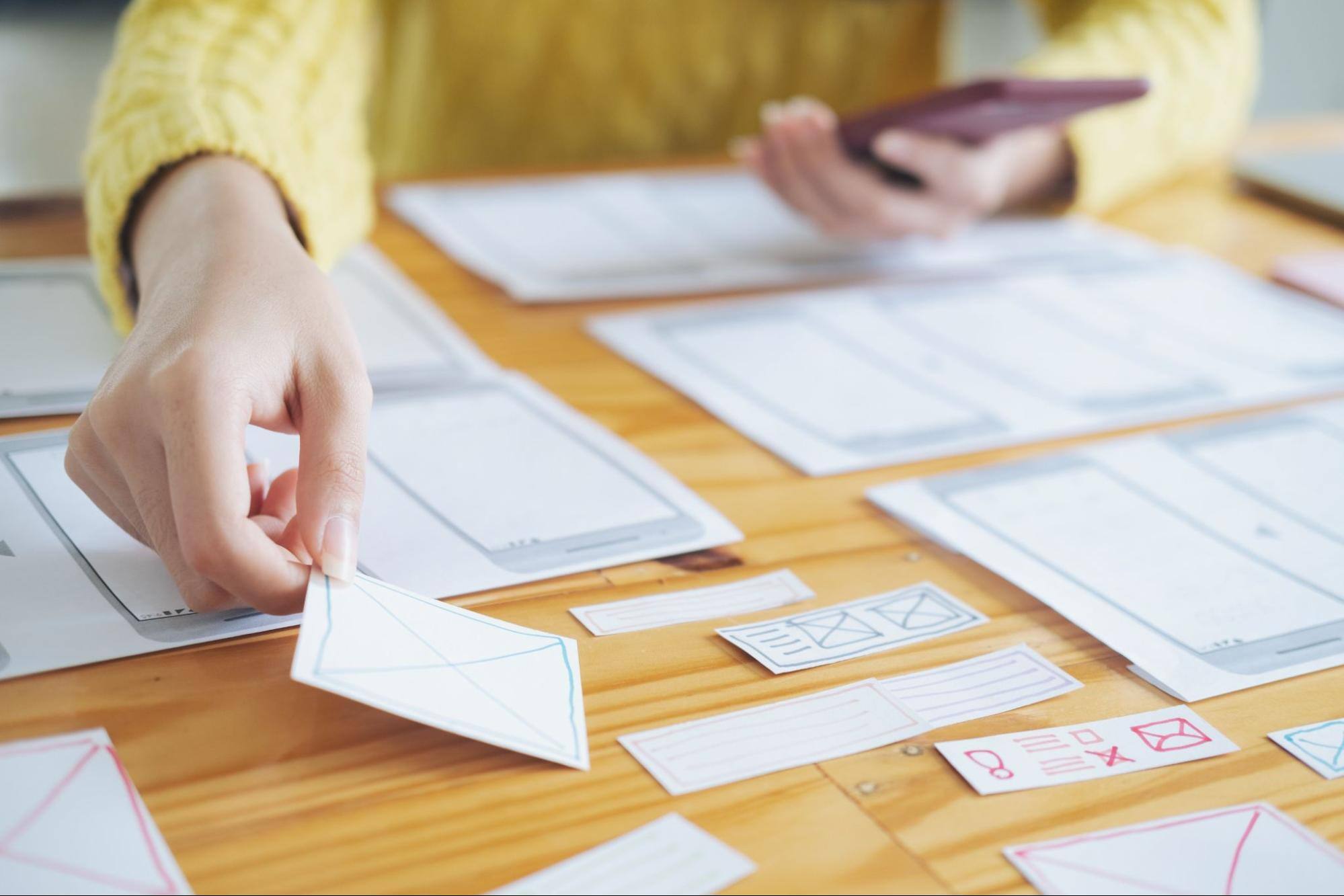 A woman in a yellow sweater arranges a layout for UX/UI and programming development technology, showcasing her hands working on a digital interface design using paper forms.